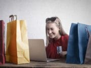 A woman is sitting at a table with multiple shopping bags, using a laptop and holding a credit card.
