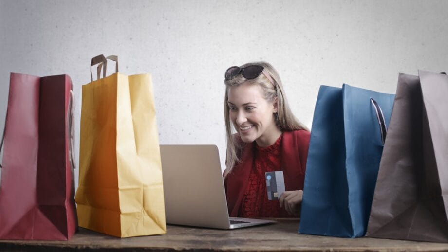 A woman is sitting at a table with multiple shopping bags, using a laptop and holding a credit card.