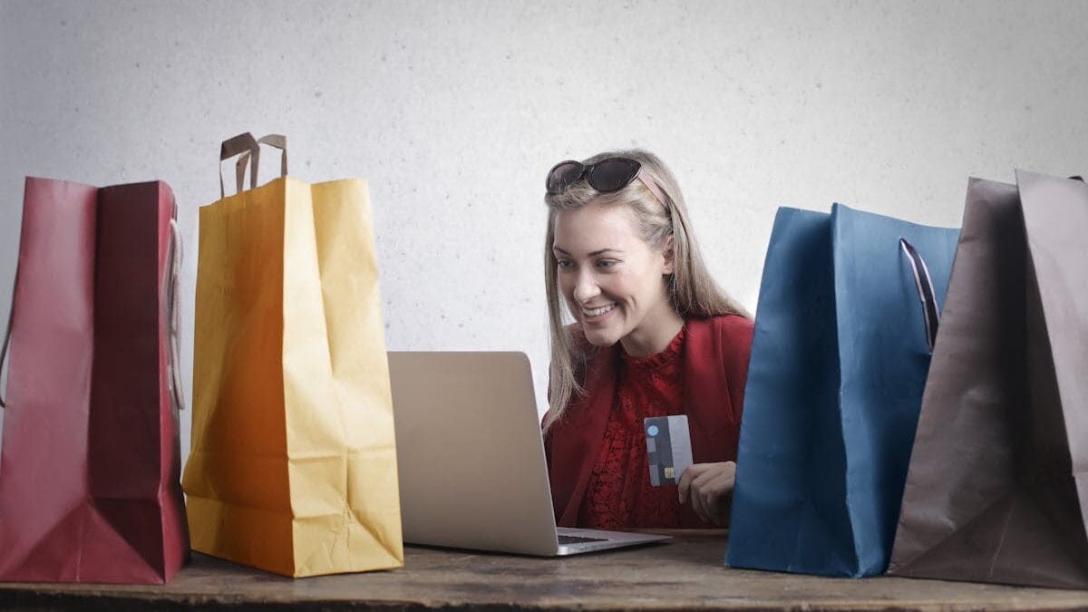 A woman is sitting at a table with multiple shopping bags, using a laptop and holding a credit card.