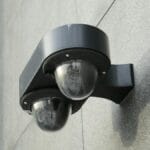 A black double dome security camera mounted on a gray concrete wall.