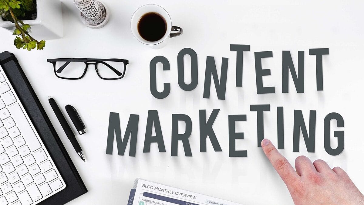 Desk with keyboard, glasses, coffee, and planner. Hand points at the words 'Content Marketing' in large letters.