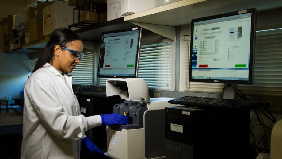 A person in a lab coat and goggles operates a piece of laboratory equipment with two computer screens displaying related data in the background.