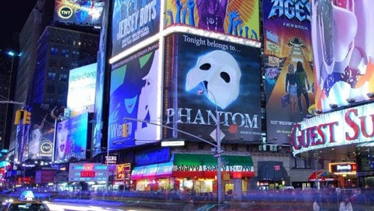 Times Square at night, featuring illuminated billboards and advertisements, including one for "Phantom." Light trails from moving cars are visible on the street below.