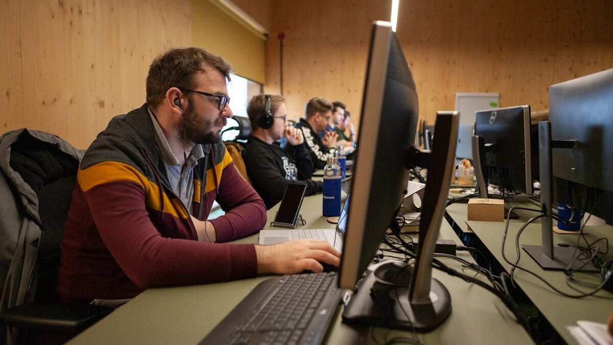 Several people are working at computers in an office setting, focusing on their screens and wearing headsets.