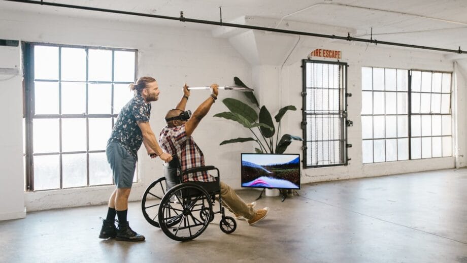 A man in a wheelchair, wearing VR goggles, raises his arms enthusiastically. Another man stands behind him, providing support. A TV screen nearby displays a racing scene.