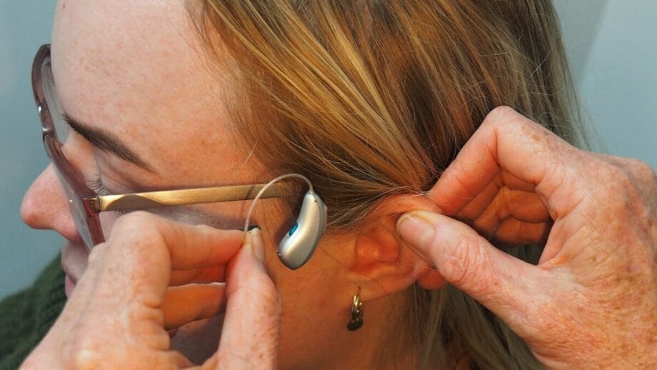 A person wearing glasses is being fitted with a hearing aid behind their ear by another individual's hands.