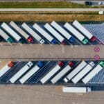 Aerial view of parked semi-trucks in angled parking spaces at a lot. A road with a few cars is visible at the top of the image.