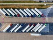 Aerial view of parked semi-trucks in angled parking spaces at a lot. A road with a few cars is visible at the top of the image.