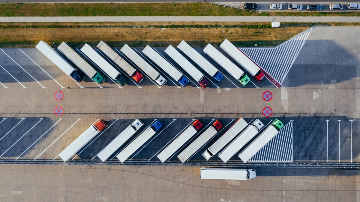 Aerial view of parked semi-trucks in angled parking spaces at a lot. A road with a few cars is visible at the top of the image.