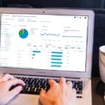 Person typing on a laptop displaying analytics with charts and graphs; a white coffee mug and pens are on the desk.