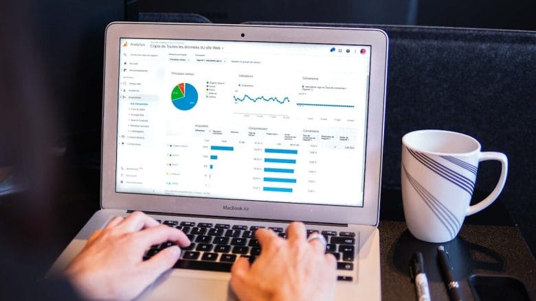 Person typing on a laptop displaying analytics with charts and graphs; a white coffee mug and pens are on the desk.