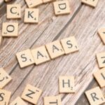 Scrabble tiles spell "SAAS" on a wooden surface, surrounded by scattered tiles with various letters.