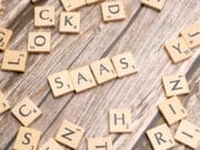 Scrabble tiles spell "SAAS" on a wooden surface, surrounded by scattered tiles with various letters.