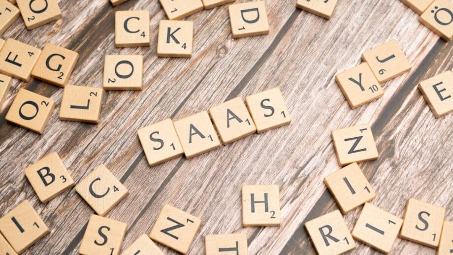 Scrabble tiles spell "SAAS" on a wooden surface, surrounded by scattered tiles with various letters.