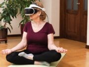 A woman sits cross-legged on a yoga mat wearing VR goggles. She has her hands resting on her knees, surrounded by indoor plants in a room with wooden flooring.
