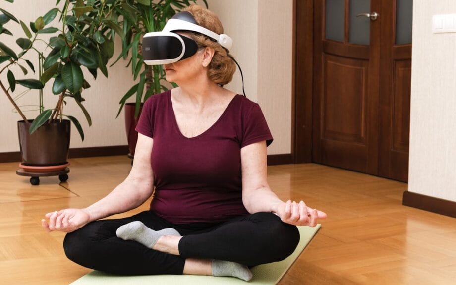 A woman sits cross-legged on a yoga mat wearing VR goggles. She has her hands resting on her knees, surrounded by indoor plants in a room with wooden flooring.