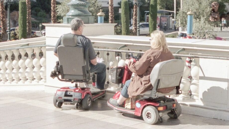 Two people on mobility scooters are stopped on a paved area beside a decorative railing with palm trees and buildings in the background.