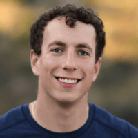 Jacob Sussman, the author of this article, with curly hair smiles at the camera, wearing a blue shirt. The background is blurred with warm tones.