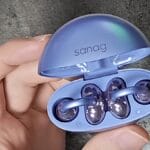 A hand holding an open blue case with two Sanag S6S Open Ear Clip Earphones inside.