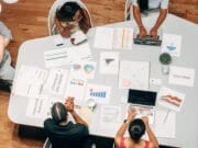 A group of five people working at a table with laptops, charts, and papers, engaging in a discussion or meeting.