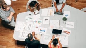 A group of five people working at a table with laptops, charts, and papers, engaging in a discussion or meeting.