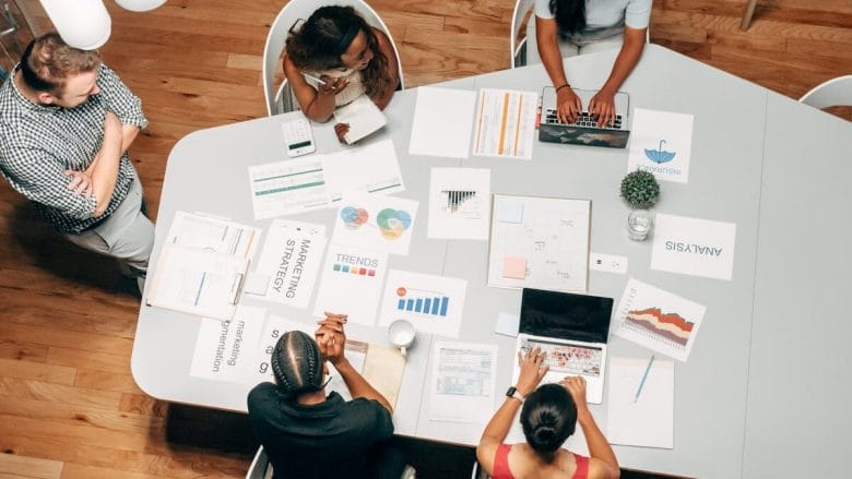 A group of five people working at a table with laptops, charts, and papers, engaging in a discussion or meeting.