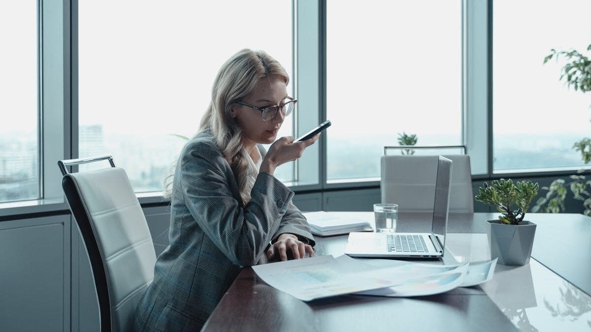 A woman in a business suit is speaking into her phone at a conference table with a laptop and documents in front of her, in a bright office.