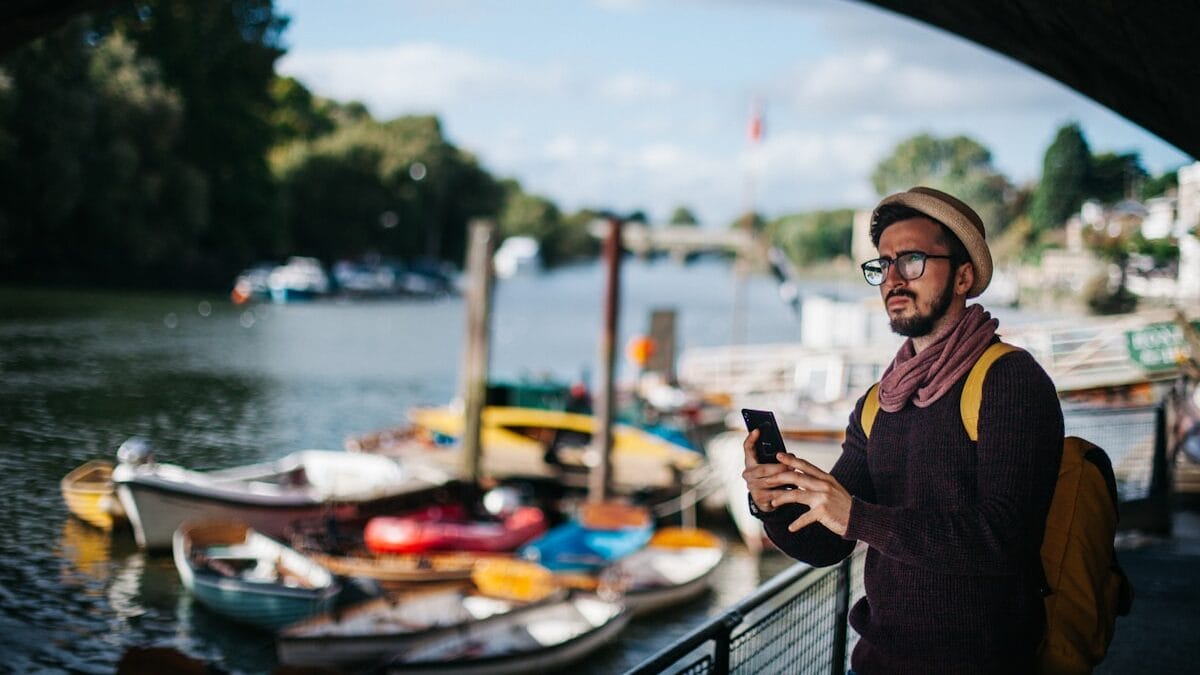 Man with hat and glasses stands under a bridge by a river, looking at his phone. Several boats are docked nearby under a partly cloudy sky.