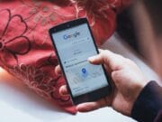 A person holding a smartphone displaying a Google Maps search with a blue location pin icon.