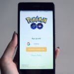 A hand holds a smartphone displaying the Pokémon GO login screen with options to sign up using Google or Pokémon Trainer Club.