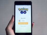 A hand holds a smartphone displaying the Pokémon GO login screen with options to sign up using Google or Pokémon Trainer Club.