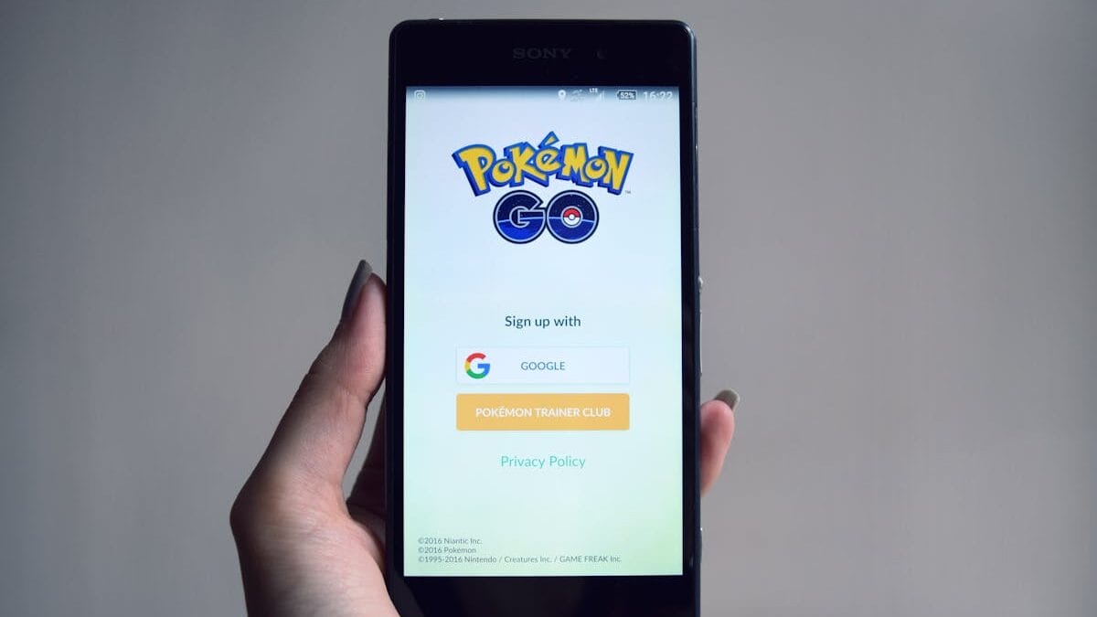 A hand holds a smartphone displaying the Pokémon GO login screen with options to sign up using Google or Pokémon Trainer Club.