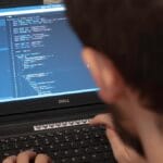 Person typing code on a Dell laptop in a cluttered workspace. Screen displays programming interface.