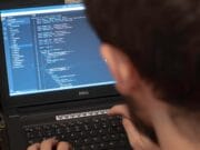 Person typing code on a Dell laptop in a cluttered workspace. Screen displays programming interface.