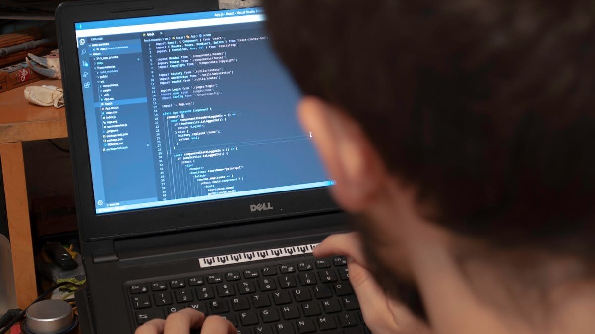 Person typing code on a Dell laptop in a cluttered workspace. Screen displays programming interface.