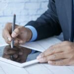 Person in a suit using a stylus on a tablet at a white desk.