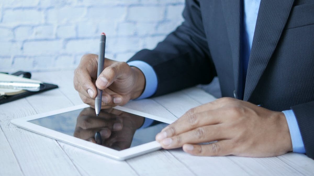 Person in a suit using a stylus on a tablet at a white desk.