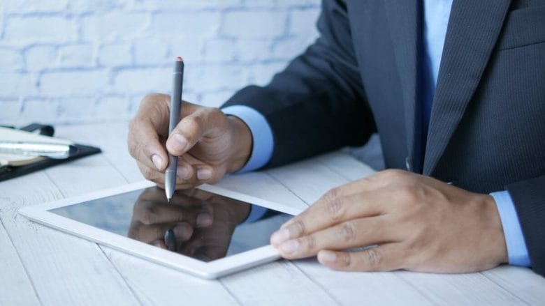 Person in a suit using a stylus on a tablet at a white desk.