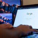 Close-up of hands typing on a laptop with the Google homepage open. A smartphone is placed on the desk, and a tablet with a mountain wallpaper is in the background.