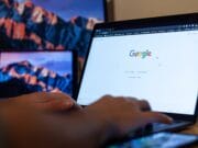 Close-up of hands typing on a laptop with the Google homepage open. A smartphone is placed on the desk, and a tablet with a mountain wallpaper is in the background.