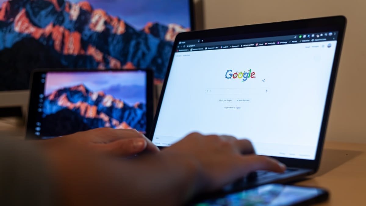 Close-up of hands typing on a laptop with the Google homepage open. A smartphone is placed on the desk, and a tablet with a mountain wallpaper is in the background.