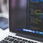 A laptop displaying a code editor with HTML and CSS code onscreen. The keyboard and part of another blurred laptop are visible in the background.