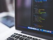 A laptop displaying a code editor with HTML and CSS code onscreen. The keyboard and part of another blurred laptop are visible in the background.