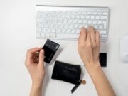 A person typing on a white keyboard holds a black credit card. A black wallet is nearby on the desk.
