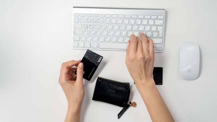 A person typing on a white keyboard holds a black credit card. A black wallet is nearby on the desk.