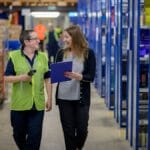 Two people walk through a warehouse aisle, one in a safety vest holding a device, and the other holding a clipboard. Shelves with various items line the aisle.