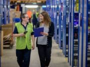 Two people walk through a warehouse aisle, one in a safety vest holding a device, and the other holding a clipboard. Shelves with various items line the aisle.