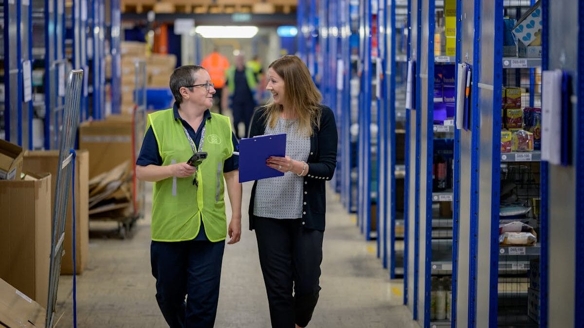 Two people walk through a warehouse aisle, one in a safety vest holding a device, and the other holding a clipboard. Shelves with various items line the aisle.