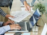 Two people are working at a desk with a laptop. One is typing, and the other is pointing at a paper with graphic designs.