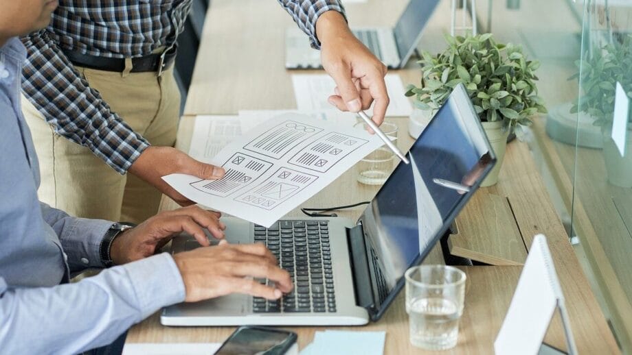 Two people are working at a desk with a laptop. One is typing, and the other is pointing at a paper with graphic designs.